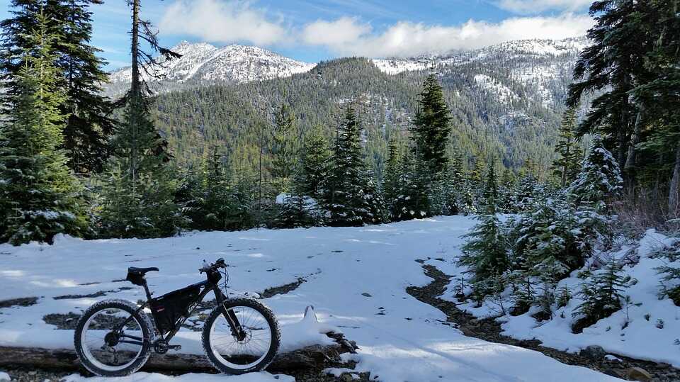 Ein Mountainbike steht an einem schnee bedeckten Berghang. Im Hintergrund sind Bäume und schnee bedeckte Berge zu sehen